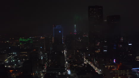 Drone-flying-over-illuminated-streets-between-tall-buildings-around-Mercantile-National-Bank-Building-with-tower-and-big-neon-clock.-Night-aerial-view-of-downtown.-Dallas,-Texas,-US