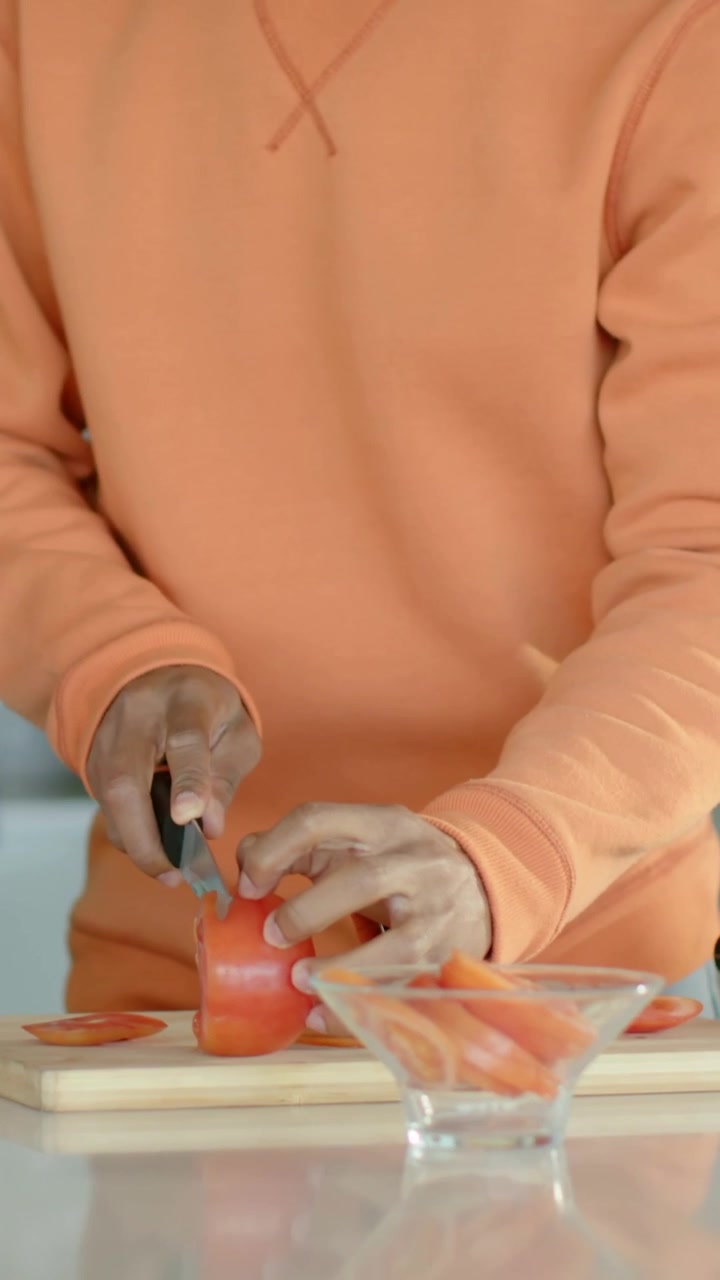 Video verticale: Entra da sinistra un uomo con una maglietta arancione che affetta un pomodoro con un coltello per la preparazione dei pasti a casa.