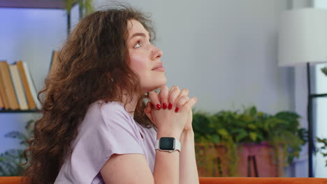 Young-woman-girl-praying-sincerely-with-folded-arms-asking-God-for-help-begging-apology-on-home-sofa