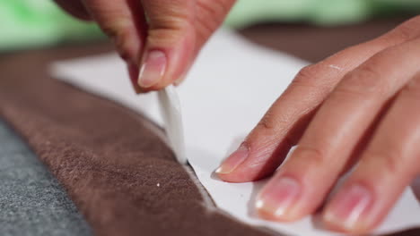 close up view of dress designer using tailors chalk to carefully trace white paper pattern on thick brown fabric during garment creation process inside professional sewing workshop
