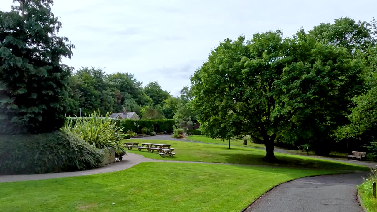 A steady camera pan reveals a lush, green botanical garden in Durham, England, with manicured lawns, mature trees, and picnic benches under soft daylight