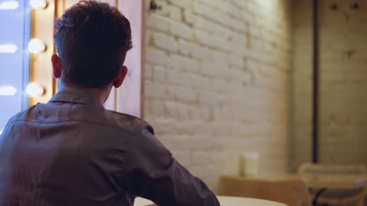 young man sitting alone at round table near window in empty cafe, coat draped over chair, back view, soft lights glowing, waiting in quiet contemplative mood amid minimal brick interior