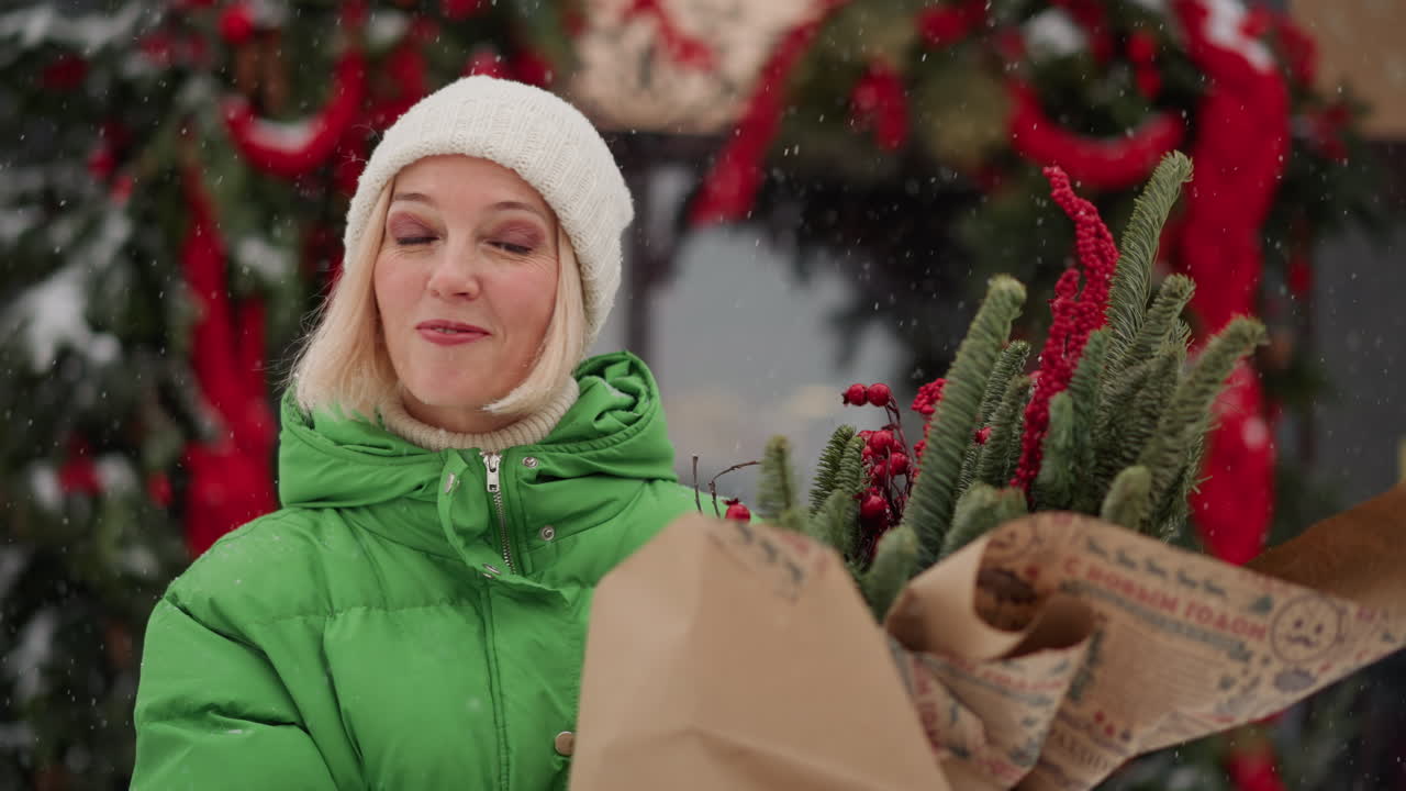 White woman carrying christmas plant bouquet, wrapped fir branches and red berries, walking past decorated storefront, gentle snowfall, green coat and knit hat, thoughtful smile, urban holiday street