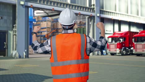 back view of a male engineer with safety helmet raising his hands celebrating while working , outside of logistics distributions warehouse