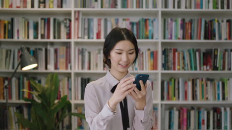 portrait-of-beautiful-asian-woman-standing-in-library-using-smartphone-texting-social-media-bookcase-in-background