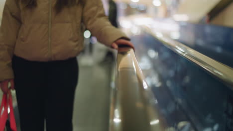 a close-up shot of a person in a peach jacket and black trousers, with their hand resting on an escalator rail. the background is blurred, emphasizing the hand's subtle grip on the shiny surface