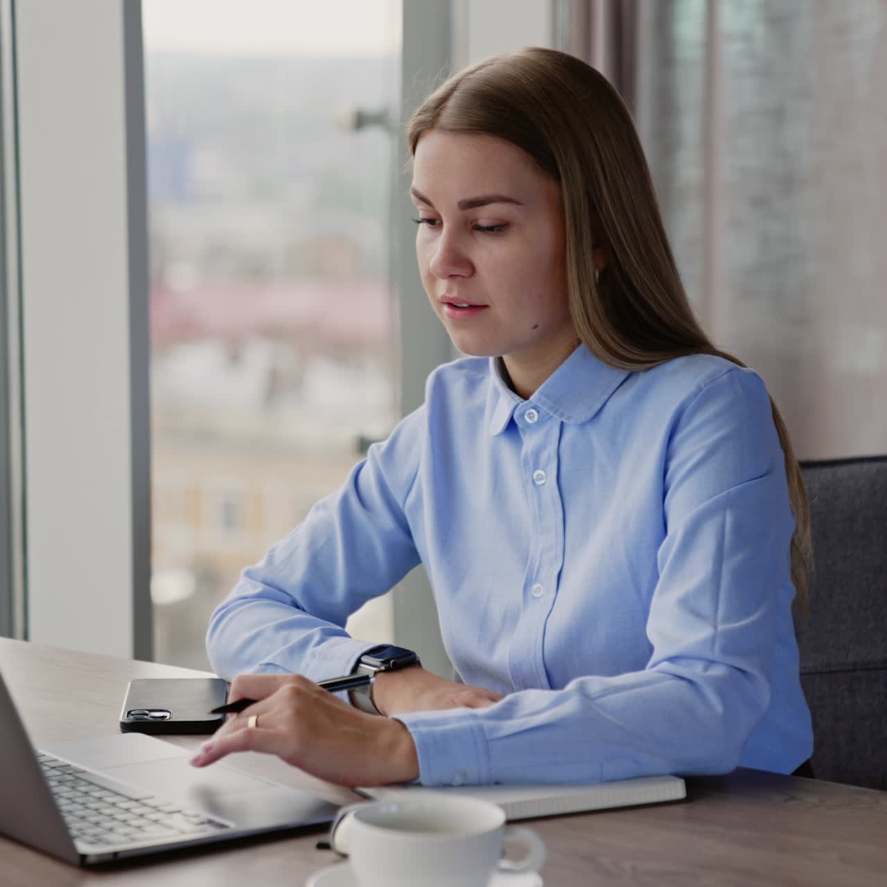 Young blonde woman taking notes into paper notebook. Lady looks at laptop in front of her and then straight ahead, thoughtfully