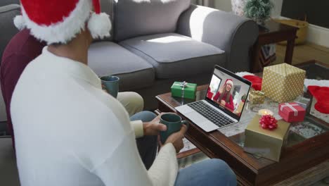 Biracial-father-and-son-with-santa-hats-using-laptop-for-christmas-video-call-with-woman-on-screen