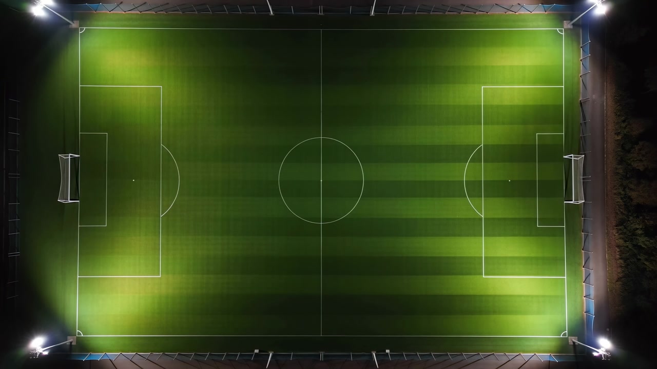 Pandangan dari atas lapangan sepak bola kosong di malam hari dengan pencahayaan sorot terang, lingkaran tengah lapangan yang terpusat, garis batas putih bersih, kotak penalti, busur sudut, rumput hijau bergaris, dan simetris