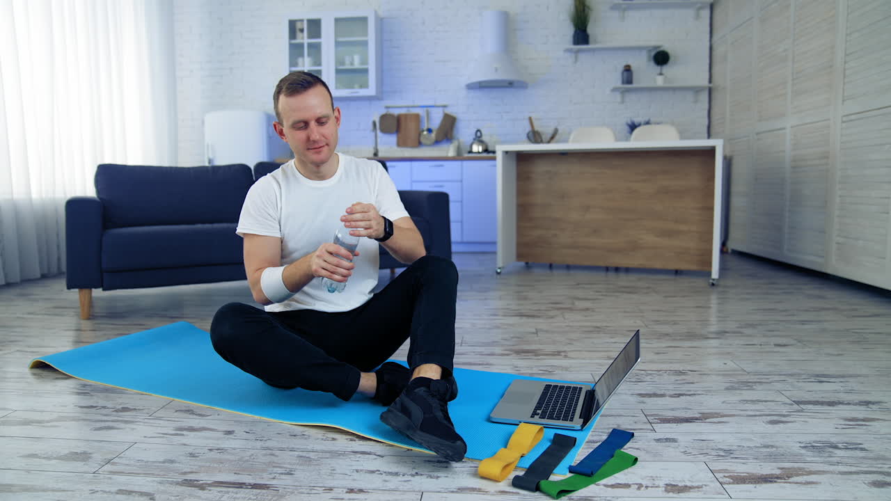 Man drinks water after daily workout at home. Guy is sitting on a fitness mat next to the laptop and holding a bottle of water. Fitness indoors. Sport during quarantine.