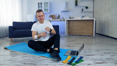 man drinks water after daily workout at home. guy is sitting on a fitness mat next to the laptop and holding a bottle of water. fitness indoors. sport during quarantine.