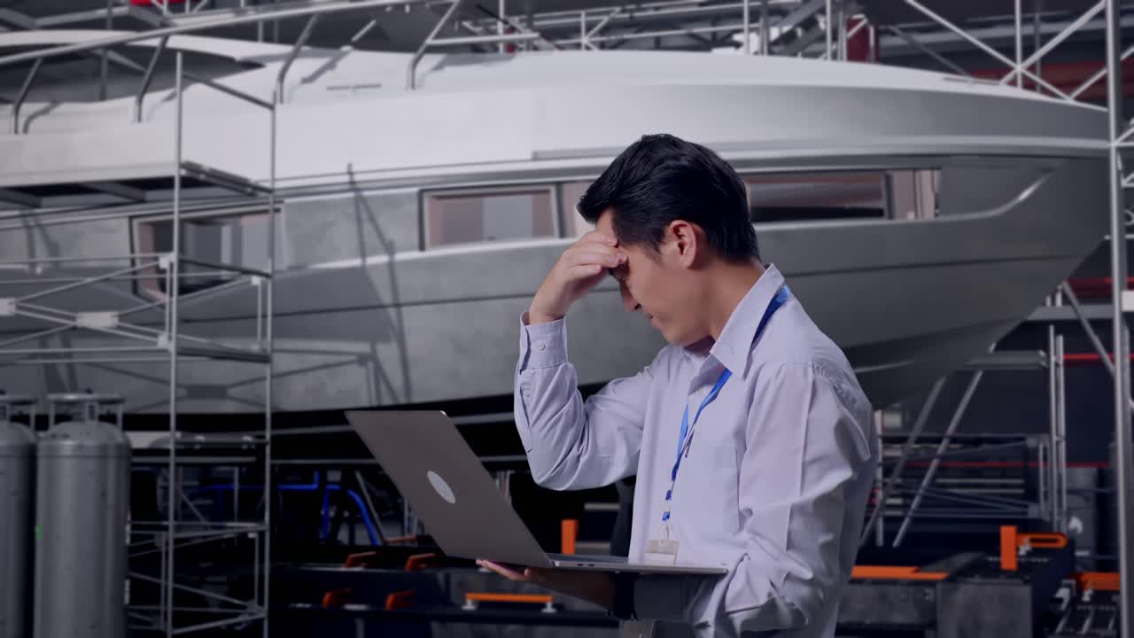 Side View Of An Asian Male Professional Worker Use Laptop with Yacht Construction in Modern Shipyard, He Is Nodding His Head With Dissapionted