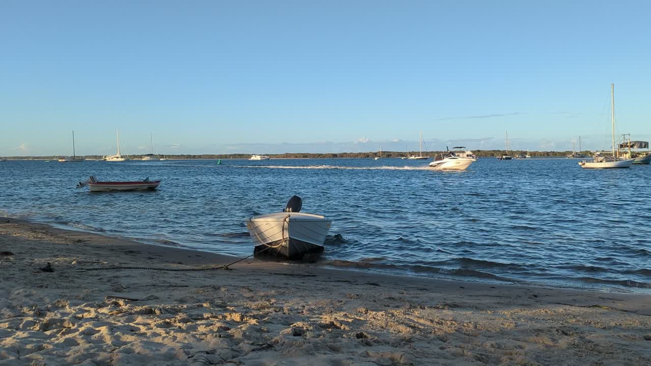 En fortøyd liten aluminiumsbåt på stranden dupper opp og ned mens en speedbåt passerer på vannet.