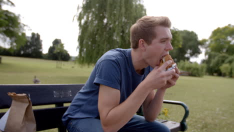 Un-Joven-Blanco-Sentado-En-Un-Banco-Del-Parque-Disfrutando-De-Una-Hamburguesa.