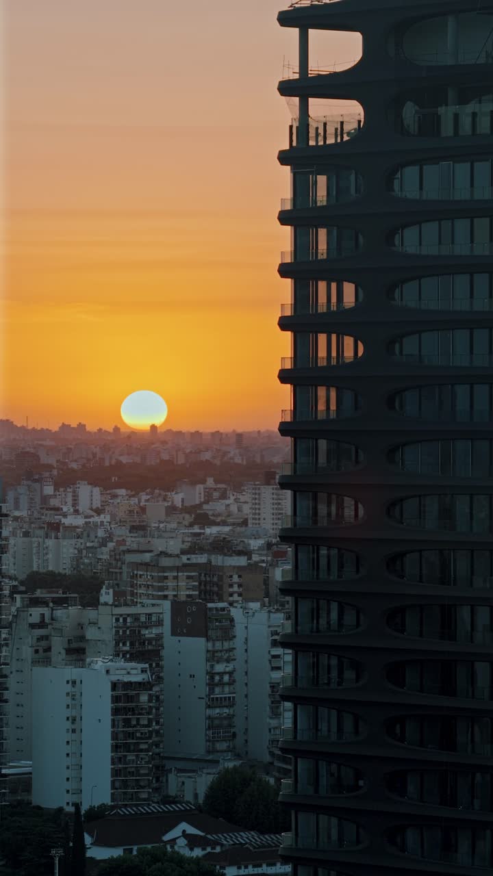Foto panorâmica vertical para a esquerda, com teleobjetiva, de um pôr do sol em Buenos Aires, apresentando uma torre residencial moderna em Palermo, luz quente e o horizonte urbano ao entardecer