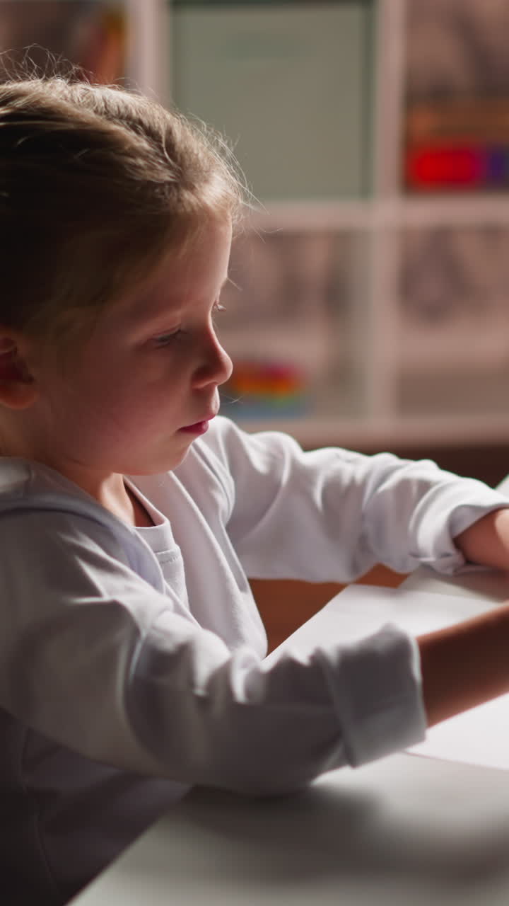 Pretty little girl in white clothes draws illustration on paper with pencil sitting at table in light children room closeup slow motion