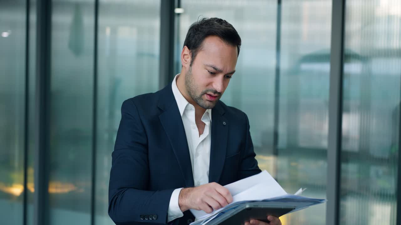 Profissional masculino estressado de terno parado em um corredor de escritório de vidro, com expressão frustrada, enquanto revisa documentos e dados financeiros negativos.