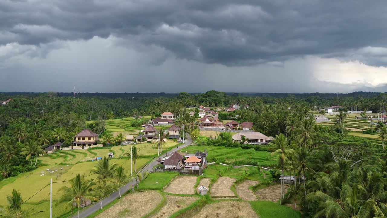 Aerial view of Bali countryside with storm clouds gathering above rice fields, houses, and dense forests, showing dramatic weather contrast with rural charm and lush green tropical landscape.