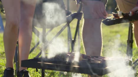 placing kebab skewers on an open campfire bbq with camp in the background