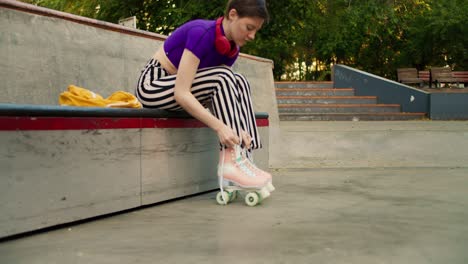 a young short-haired girl in a purple top and striped pants laces up roller skates on a skateboarding site in the park in summer