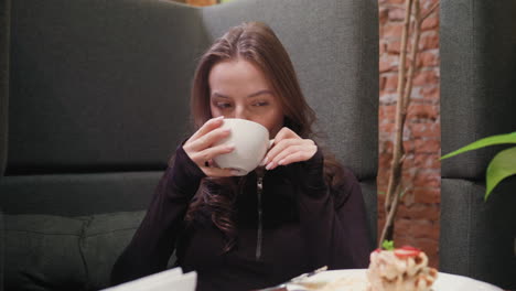 lady sits relaxed in cozy booth with dark upholstery and exposed brick wall, holding white ceramic cup of latte while waffles rest on table beside vase with green plant, evoking serene atmosphere