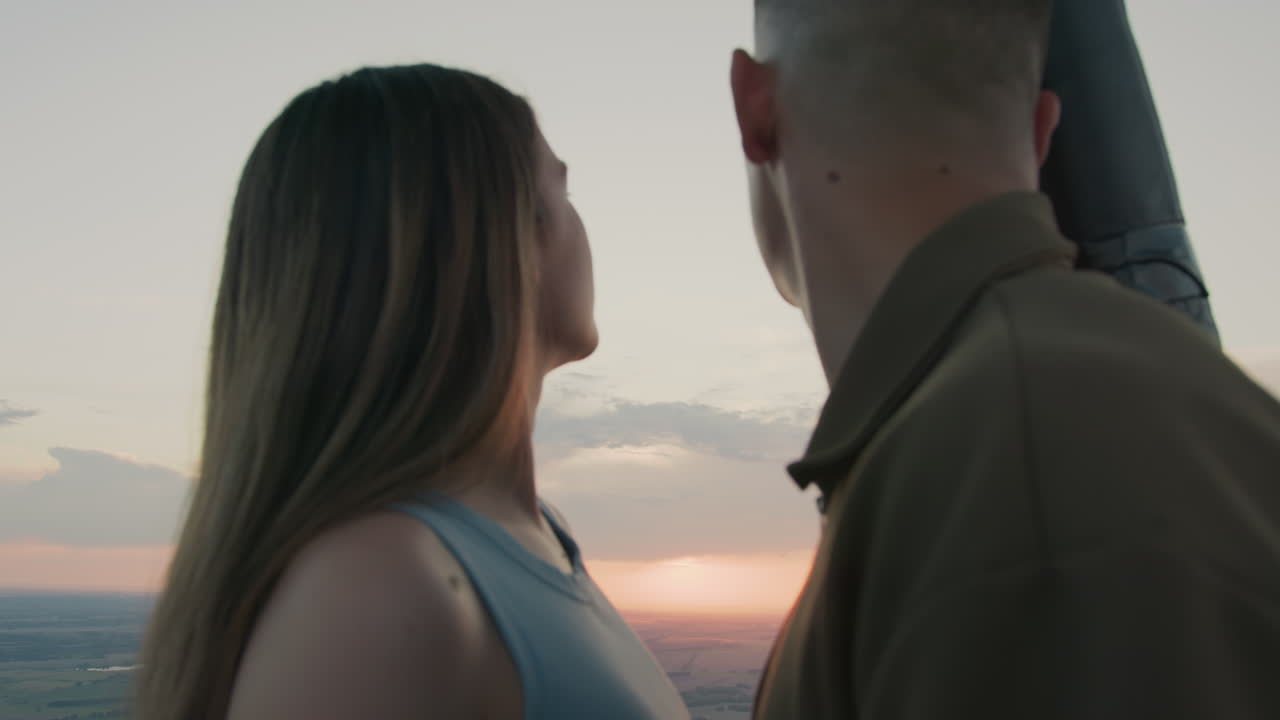 Close up of couple enjoying sunset gaze as distant red paraglider drifts across pastel sky above sprawling farmland under soft clouds creating serene romantic aerial adventure moment