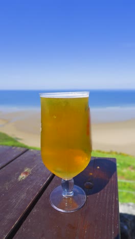 vertical video freshly poured chilled lager beer on wooden table overlooking beautiful beach landscape with bright blue sky. refreshing summer alcoholic drink.