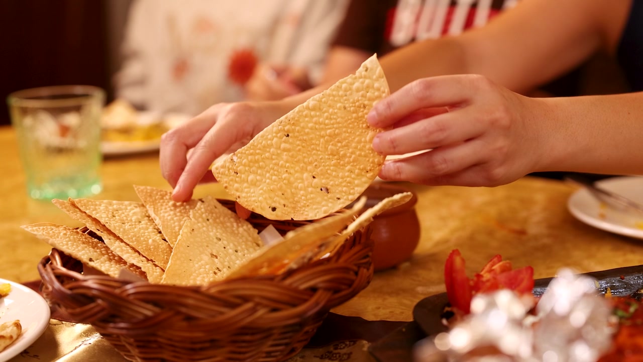 Primo piano di commensali che prendono pane papadum croccante da un cestino su un tavolo di ristorante.