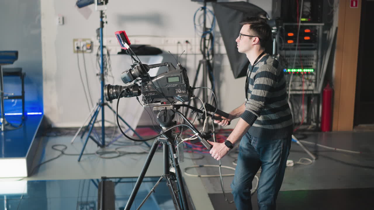 Man adjusting professional video camera on tripod in studio setting. Filming session in progress with equipment setup and cables visible. Technician operating camera during production