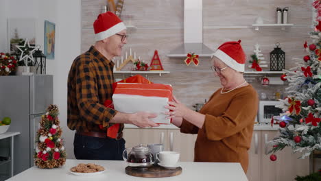 Pareja-Mayor-Con-Sombrero-De-Claus-Sanata-Compartiendo-Regalo-De-Envoltorio-De-Navidad-Con-Cinta