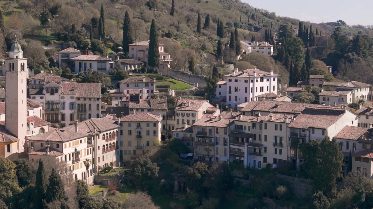 Storica città collinare con edifici in pietra e torre circondata da una vegetazione lussureggiante.