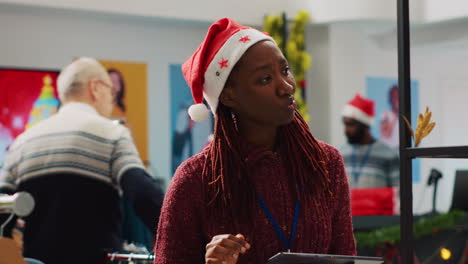 Employee-wearing-Santa-hat-inputing-price-list-on-tablet,-adding-them-to-online-shop-pages.-Retail-assistant-working-in-Christmas-decorated-clothing-store-during-winter-holiday-season