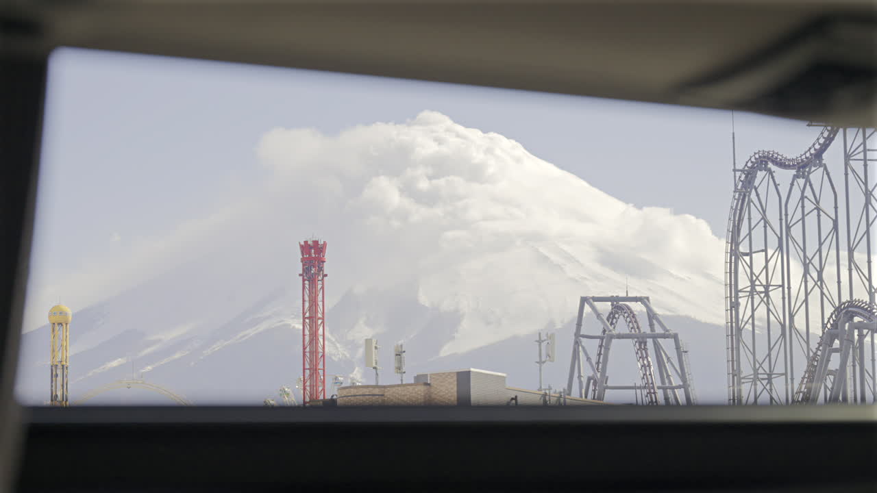 A stunning landscape shows a snow-covered Fuji mountain in the background, contrasting with colorful amusement park rides in the foreground on a clear, sunny day