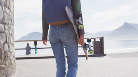 Rückansicht,-Mittelteil-Einer-Frau-Mit-Gemischter-Abstammung,-Die-Skateboard-Hält-Und-Auf-Der-Promenade-Am-Meer-Spaziert