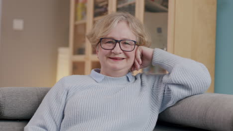 Smiling-elderly-caucasian-woman-with-glasses-sits-relaxed-on-sofa,-frontal