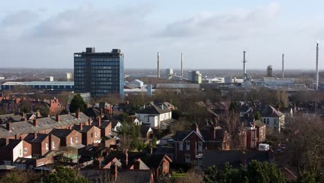 Aerial-view-over-park-trees-to-urban-industrial-townscape-with-blue-skyscraper,-Merseyside,-England