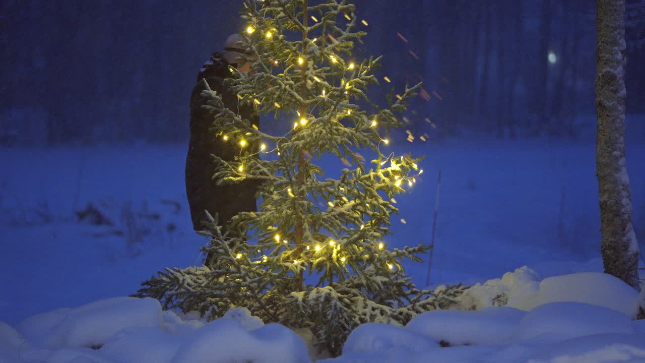 Nainen koristelee joulukuusta hämärässä lumisessa puutarhassaan.