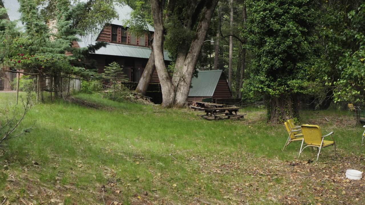 Flying low towards an old wooden cabin with weathered picnic tables in garden, abandoned lodge in forest, aerial