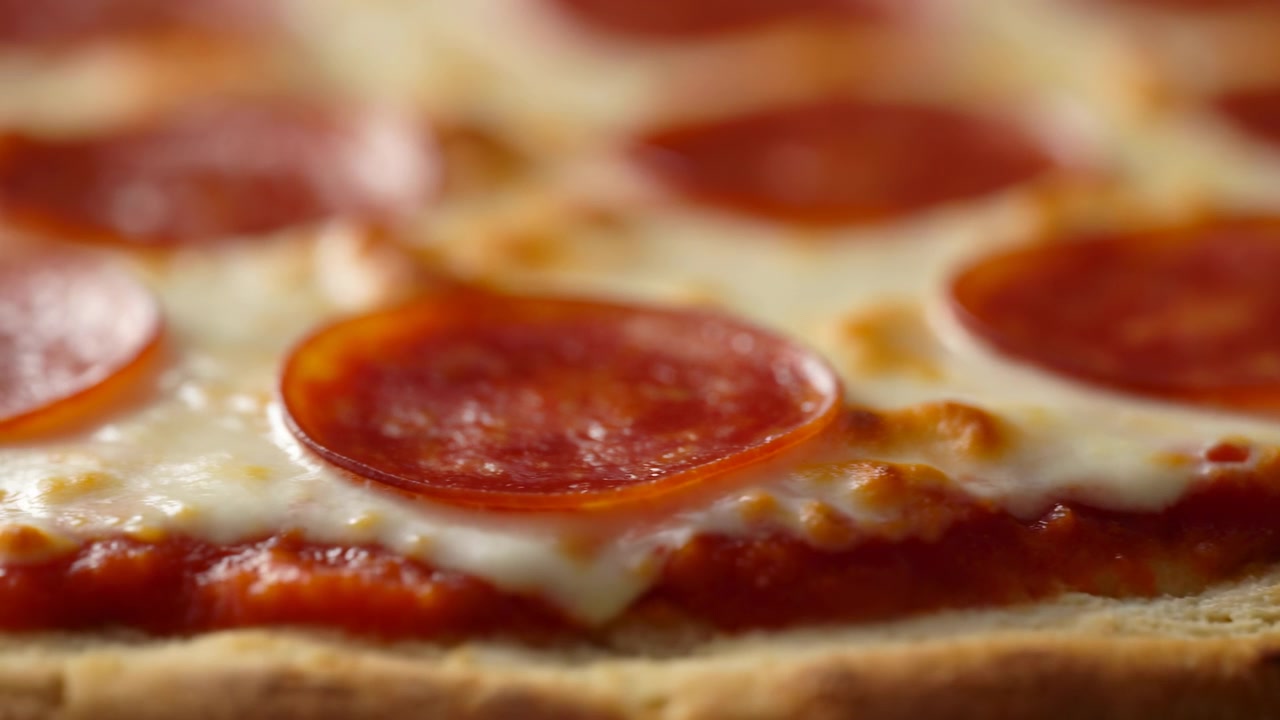 Yavaş yanal çerçeveleme, erimiş peynir ve kırmızı domates sosu üzerinde parlak pepperoni dilimlerini, alt kenar boyunca kızarmış kabuğu ve arkadaki malzemeleri yumuşatan sığ odaklamayı gösteriyor