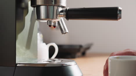 las gotas de bebida caen sobre la bandeja de la cafetera. una mujer joven sostiene una taza de cerámica de café listo cerca de la máquina de café. la comodidad de la rutina matutina
