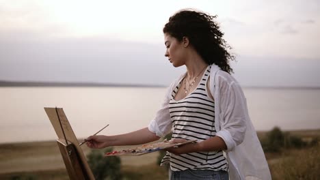 side view of an attractive curly woman artist working on her picture with easel outdoors near the lake. morning, soft wind blowing on woman's face