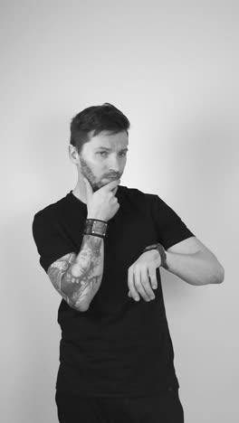 grayscale footage of a white tattooed man with short hair and beard looks at his wristwatch and begins tapping it while looking into the camera, showing your running out of time soon, hurry please.