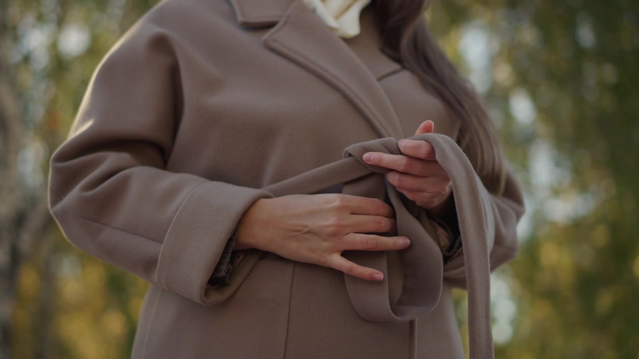 Primo piano di una donna incinta che si sistema la cintura del cappotto marrone in un ambiente esterno con luce naturale soffusa. Le sue mani sono visibili mentre manipola delicatamente il tessuto, mettendo in mostra texture e movimento.