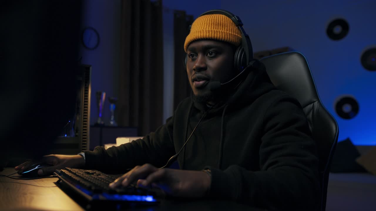 Male professional computer gamer looking at computer screen, talks to team through headset, presses buttons of backlit keyboard. Dark room lit by blue led light
