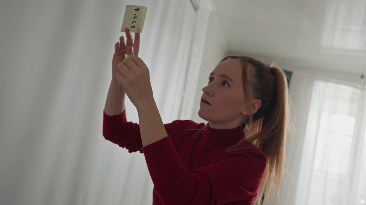 Young woman in red turtleneck examines and places sticky. Woman adjusts note positions with both hands focusing intently. Bright room features sheer curtains dark green drapes