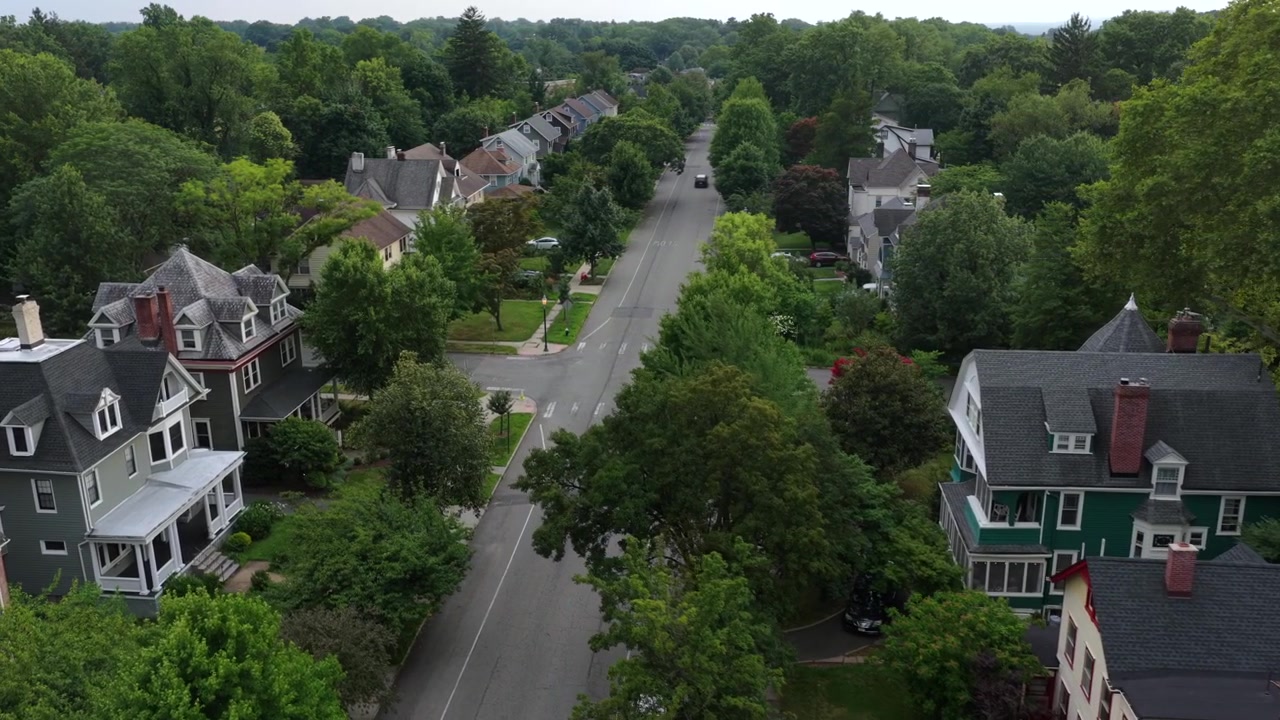 Victorian style houses with turrets line tree shaded street near Seton Hall in South Orange, New Jersey, dense green canopy covers quiet residential neighborhood streets. Aerial drone flyover shot