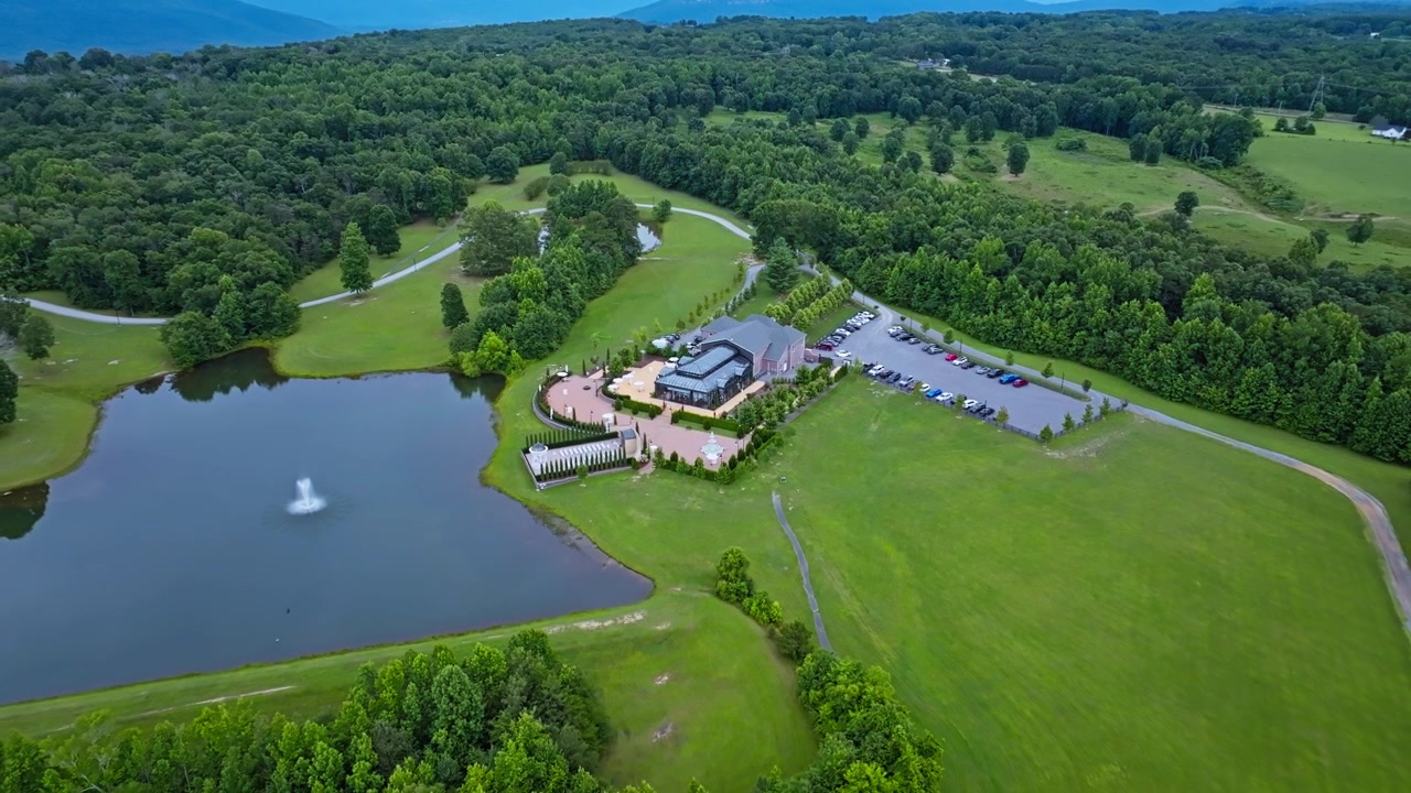 Elegant bryllupslokale med glastag og buet stenbelagt terrasse ligger ved en rolig dam nær Trenton, Georgia. En overfyldt parkeringsplads grænser op til tæt grøn sommer-skov. Luftfoto fra drone, top-down