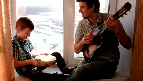 playing a musical instrument. dad is playing the guitar and son is playing tambourine sitting in windowsill.