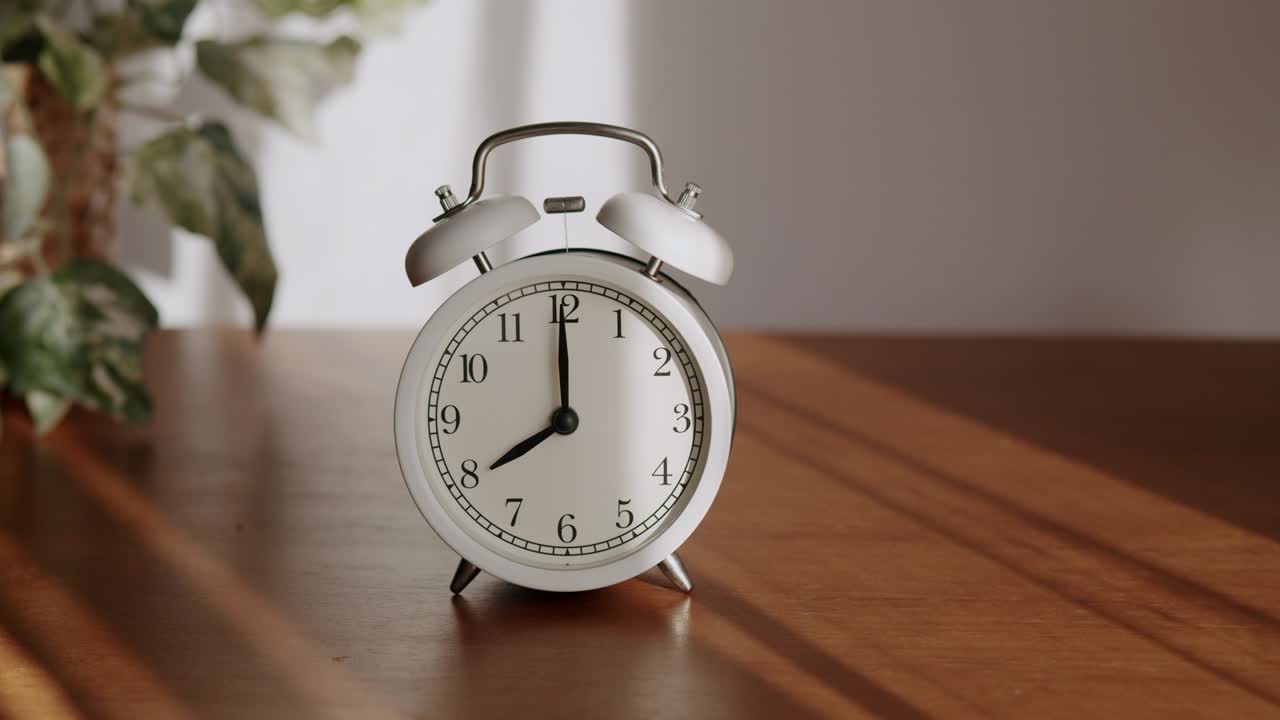 White alarm clock is placed on a wooden table and the light shines through the curtains in the morning