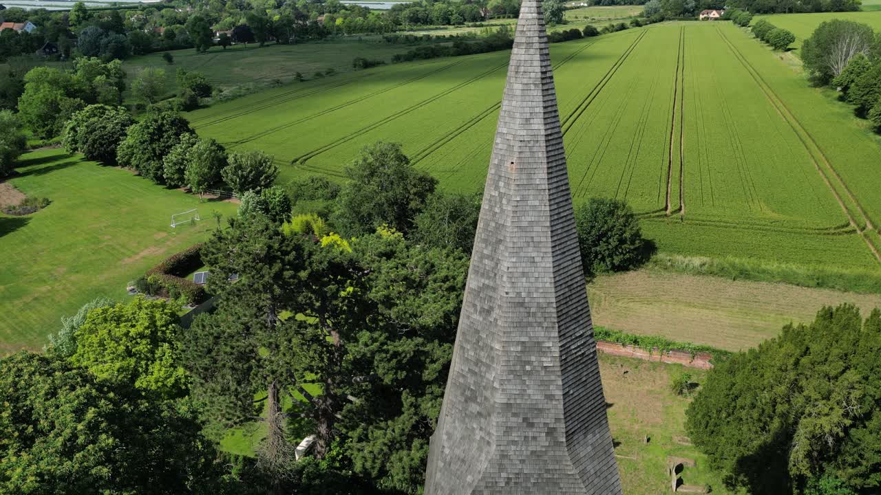 Rising boom shot of St John the Evangelist church's spire, in Ickham, Kent, with a field and graveyard in the background